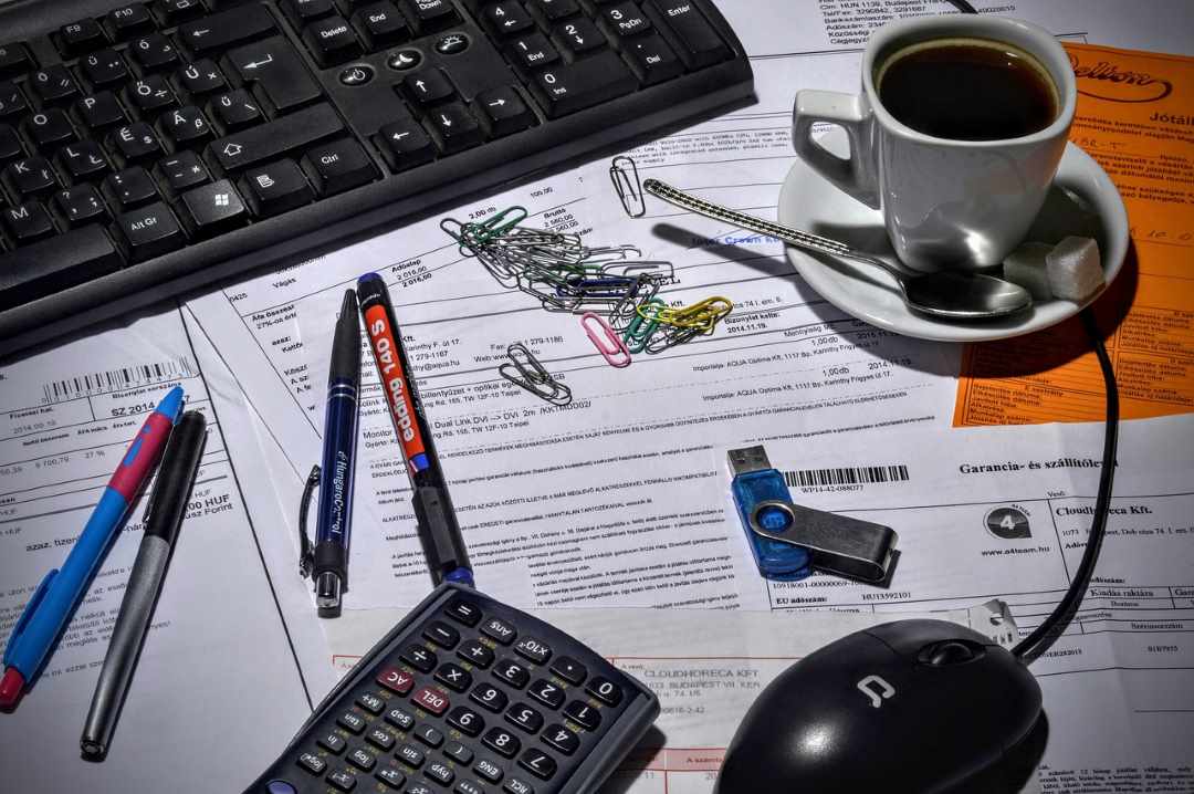 En una mesa boligrafos, pendrive, cafe, calculadora, ratón, ordenador y clips